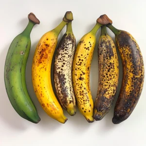 An image illustrating the stages of banana ripening, transitioning from green bananas to yellow and finally to brown or black overripe bananas perfect for baking. MidJourney Prompt: Five bananas displayed side-by-side, showing gradual ripeness stages — green, yellow, yellow with spots, brown-spotted, and fully black peel — against a white background, clear and educational presentation.