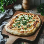 A freshly baked flatbread topped with melted cheese and chopped herbs, served on a wooden board with a rustic background.