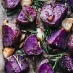 A close-up of roasted purple potatoes, golden garlic cloves, and fresh rosemary sprigs on a baking sheet, seasoned with herbs and spices for a delicious and flavorful dish.