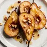 Caramelized baked pear halves drizzled with honey and topped with crunchy nuts on a speckled white plate