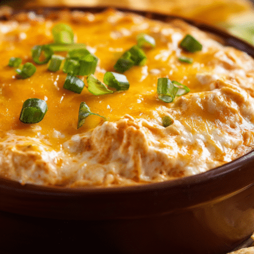 Close-up of hot buffalo chicken dip in a brown baking dish, topped with melted cheddar cheese and chopped green onions, with tortilla chips on the side for dipping.